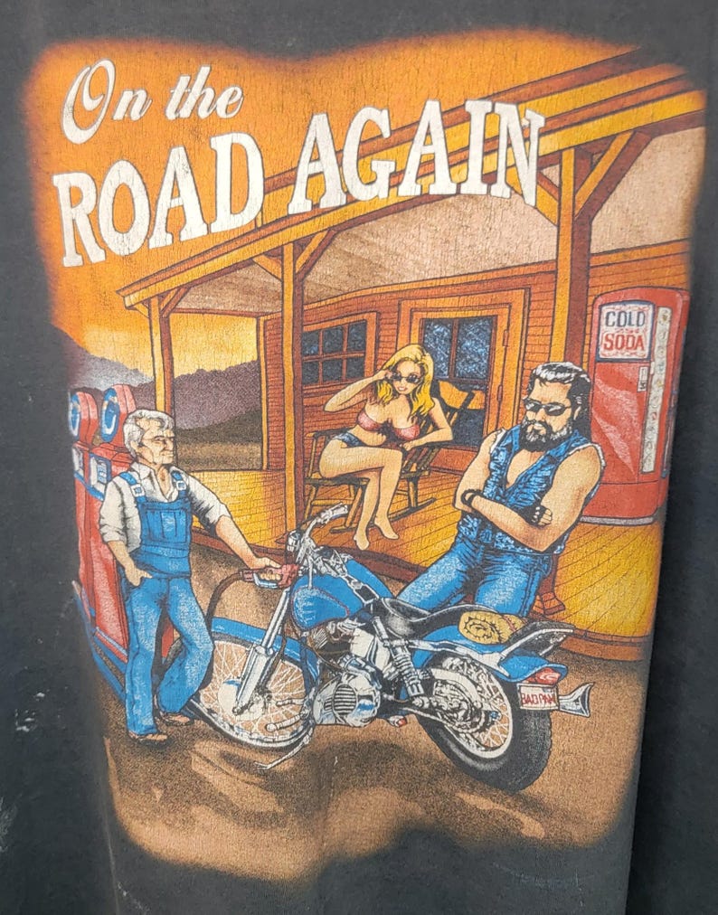 Peut inclure: T-shirt noir avec un motif repr&eacute;sentant une moto, une station-service et trois personnages. Le texte "On the Road Again" est imprim&eacute; en haut. L'image a un style vintage et r&eacute;tro.