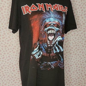 K&ouml;nnte beinhalten: Schwarzes T-Shirt mit dem Bandnamen "Iron Maiden" in roten und wei&szlig;en Buchstaben. Die Grafik zeigt eine skelettartige Figur mit blauen und roten Details, die eine Brille und Kopfh&ouml;rer tr&auml;gt, mit offenem Mund und ausgestreckten H&auml;nden.