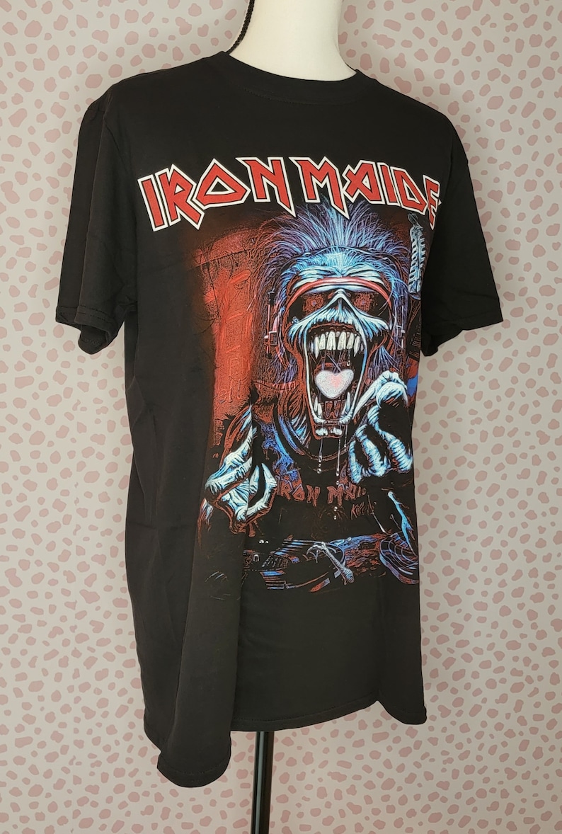 K&ouml;nnte beinhalten: Schwarzes T-Shirt mit dem Bandnamen "Iron Maiden" in roten und wei&szlig;en Buchstaben. Die Grafik zeigt eine skelettartige Figur mit blauen und roten Details, die eine Brille und Kopfh&ouml;rer tr&auml;gt, mit offenem Mund und ausgestreckten H&auml;nden.