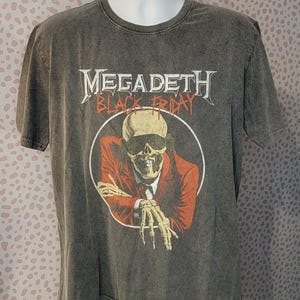 May include: Gray t-shirt featuring the band name "Megadeth" in white, above a graphic of a skeleton in a red suit and sunglasses. The words "Black Friday" are below the band name. The shirt has a crew neck and short sleeves.