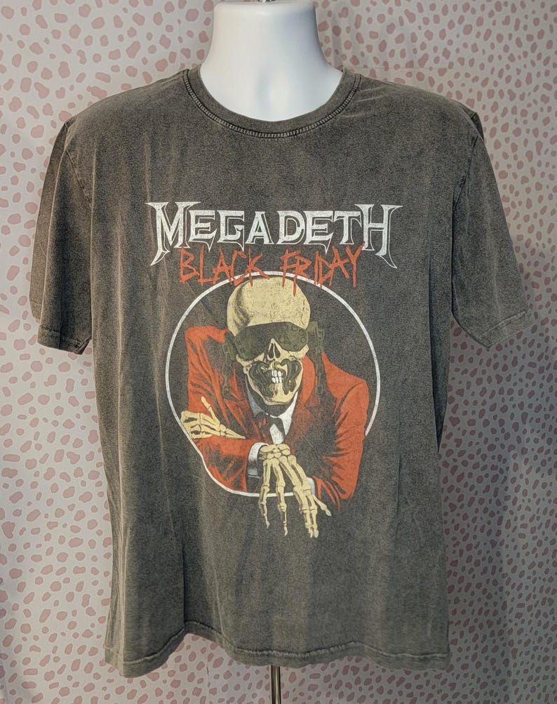 May include: Gray t-shirt featuring the band name "Megadeth" in white, above a graphic of a skeleton in a red suit and sunglasses. The words "Black Friday" are below the band name. The shirt has a crew neck and short sleeves.