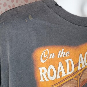 Peut inclure: T-shirt gris fonc&eacute; avec l'inscription "On the Road Again" en blanc et orange. Le graphisme repr&eacute;sente une sc&egrave;ne de coucher de soleil avec un b&acirc;timent et un v&eacute;hicule. Le t-shirt a un col rond et des manches courtes.