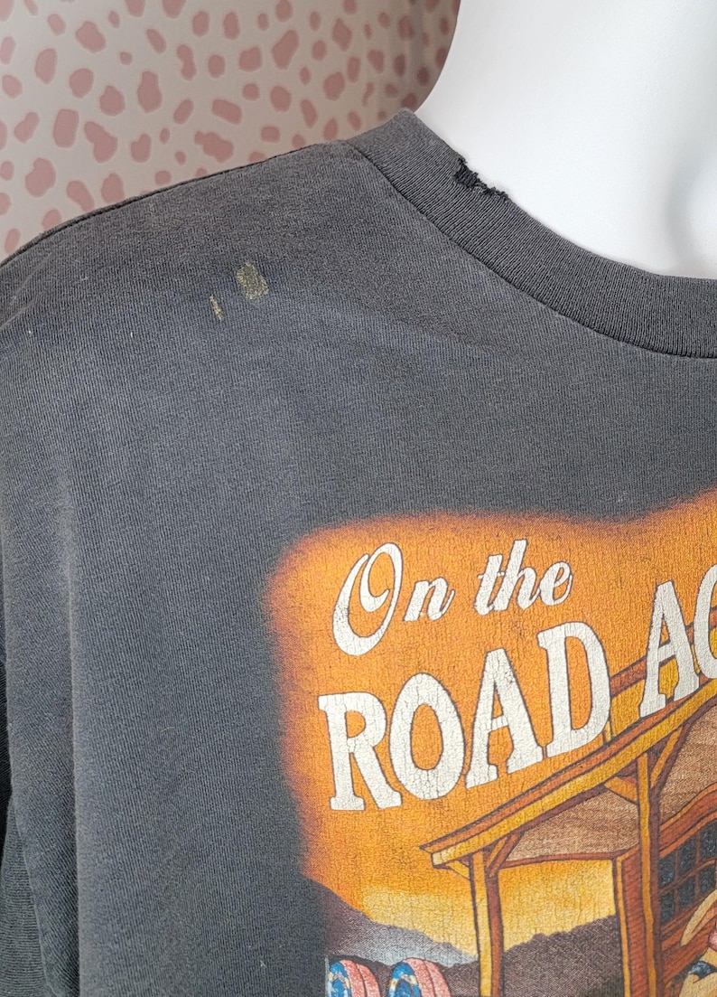 Peut inclure: T-shirt gris fonc&eacute; avec l'inscription "On the Road Again" en blanc et orange. Le graphisme repr&eacute;sente une sc&egrave;ne de coucher de soleil avec un b&acirc;timent et un v&eacute;hicule. Le t-shirt a un col rond et des manches courtes.