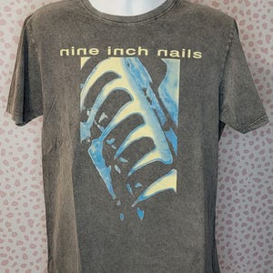 May include: A gray Nine Inch Nails t-shirt. The shirt features the band's name in yellow block letters above a blue and yellow graphic design. The shirt is displayed on a mannequin.