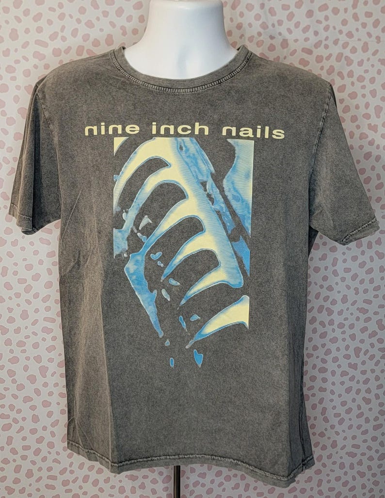 May include: A gray Nine Inch Nails t-shirt. The shirt features the band's name in yellow block letters above a blue and yellow graphic design. The shirt is displayed on a mannequin.