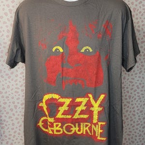 May include: Gray t-shirt featuring a red and yellow graphic of a face and the name "Ozzy Osbourne." The graphic is distressed, giving it a vintage look. The shirt has short sleeves and a crew neck.