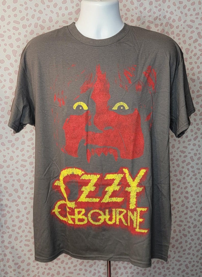 May include: Gray t-shirt featuring a red and yellow graphic of a face and the name "Ozzy Osbourne." The graphic is distressed, giving it a vintage look. The shirt has short sleeves and a crew neck.