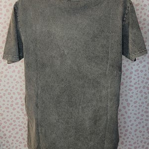 May include: A gray, short-sleeved t-shirt with a textured, washed appearance. The shirt is displayed on a mannequin. The fabric has a mottled effect, with variations in the gray color. The neckline and sleeves are finished with a simple hem.