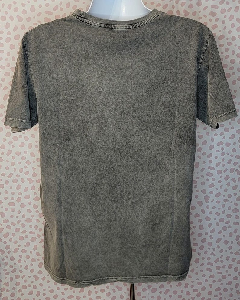 May include: A gray, short-sleeved t-shirt with a textured, washed appearance. The shirt is displayed on a mannequin. The fabric has a mottled effect, with variations in the gray color. The neckline and sleeves are finished with a simple hem.