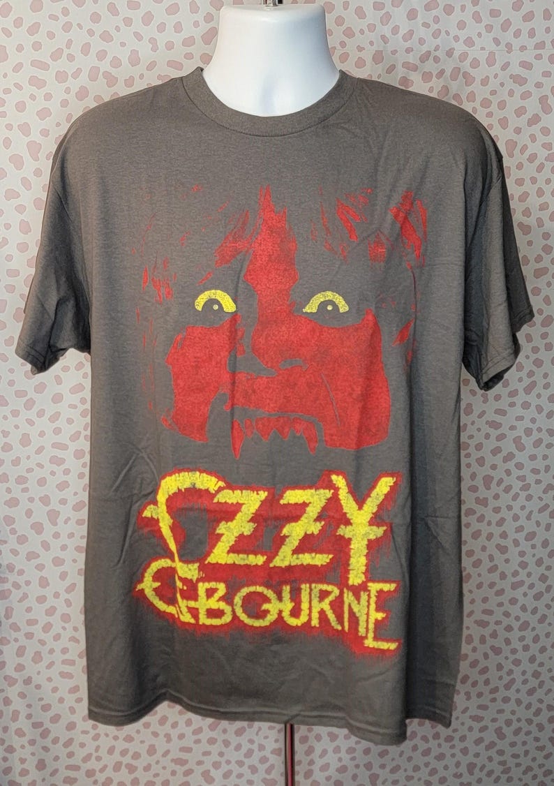 May include: A gray t-shirt featuring a red graphic of a face with yellow eyes and the name "OZZY OSBOURNE" in yellow with a red outline. The shirt is displayed on a mannequin.