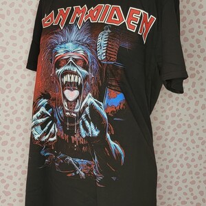K&ouml;nnte beinhalten: Schwarzes T-Shirt mit dem Bandnamen "Iron Maiden" in roter Schrift. Die Grafik zeigt ein Monster mit Mikrofon in Rot, Blau und Wei&szlig;. Das T-Shirt wird auf einer Schaufensterpuppe pr&auml;sentiert.