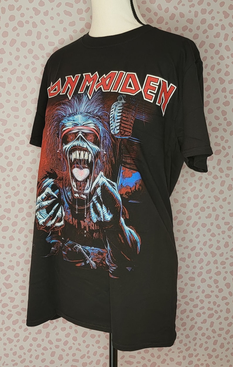 K&ouml;nnte beinhalten: Schwarzes T-Shirt mit dem Bandnamen "Iron Maiden" in roter Schrift. Die Grafik zeigt ein Monster mit Mikrofon in Rot, Blau und Wei&szlig;. Das T-Shirt wird auf einer Schaufensterpuppe pr&auml;sentiert.