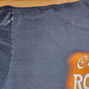 Peut inclure: T-shirt bleu marine d&eacute;lav&eacute; avec l'inscription "On the Road Again" en blanc et orange. Le t-shirt a un col rond et des manches courtes. Le tissu montre des signes d'usure.