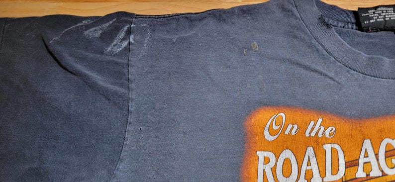 Peut inclure: T-shirt bleu marine d&eacute;lav&eacute; avec l'inscription "On the Road Again" en blanc et orange. Le t-shirt a un col rond et des manches courtes. Le tissu montre des signes d'usure.