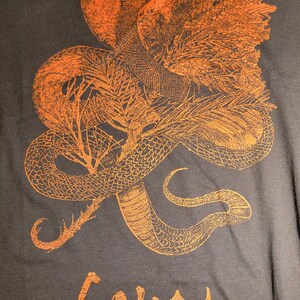K&ouml;nnte beinhalten: Ein dunkelgraues T-Shirt mit einem orangefarbenen Druck eines Schlangen, das sich um einen Baum mit einer Sonne dahinter windet. Der Text "GOJIRA" ist in Orange auf der Unterseite des T-Shirts gedruckt.
