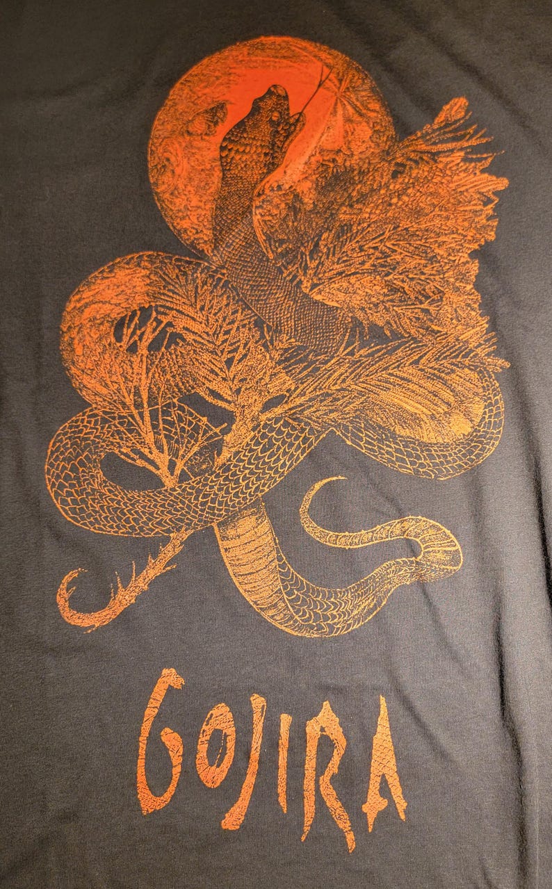 K&ouml;nnte beinhalten: Ein dunkelgraues T-Shirt mit einem orangefarbenen Druck eines Schlangen, das sich um einen Baum mit einer Sonne dahinter windet. Der Text "GOJIRA" ist in Orange auf der Unterseite des T-Shirts gedruckt.