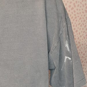 Peut inclure: Un t-shirt gris d&eacute;lav&eacute; &agrave; manches courtes. Le t-shirt a un aspect l&eacute;g&egrave;rement us&eacute;, avec quelques marques de peinture blanche sur la manche. Le tissu semble doux et us&eacute;. Le fond est un motif &agrave; pois roses et blancs.