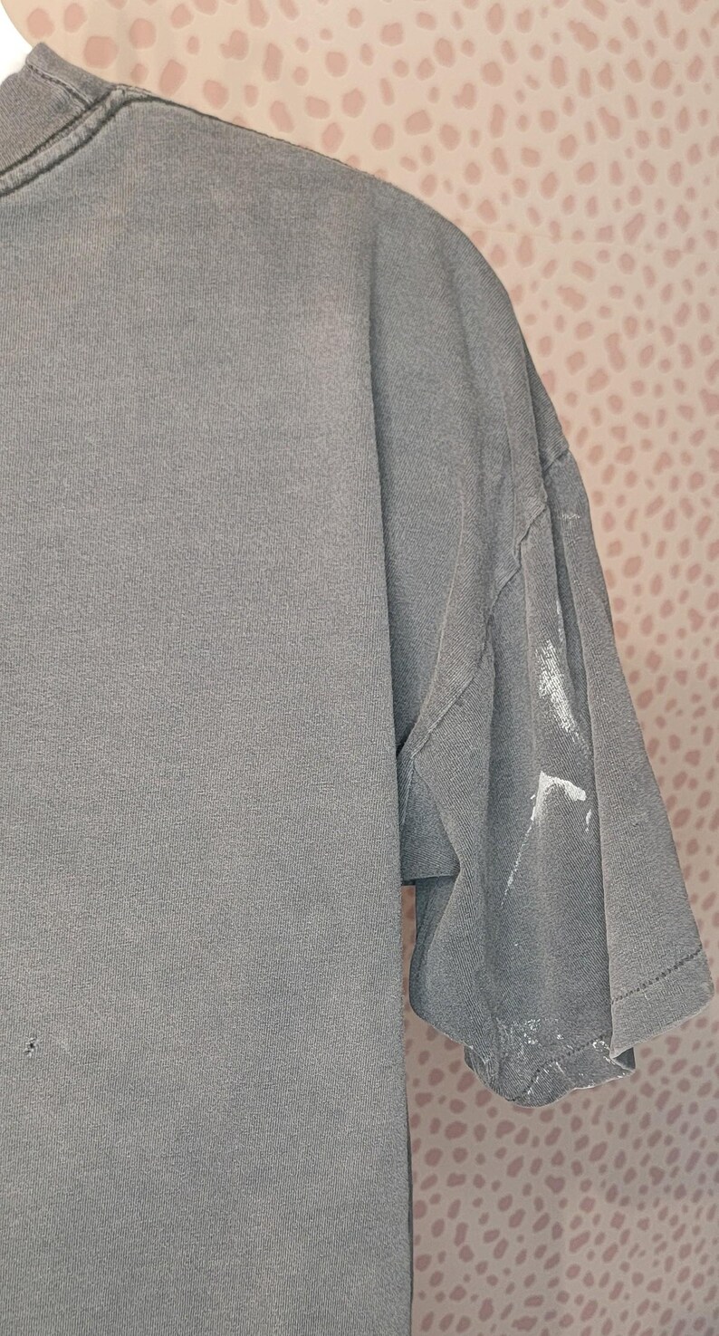 Peut inclure: Un t-shirt gris d&eacute;lav&eacute; &agrave; manches courtes. Le t-shirt a un aspect l&eacute;g&egrave;rement us&eacute;, avec quelques marques de peinture blanche sur la manche. Le tissu semble doux et us&eacute;. Le fond est un motif &agrave; pois roses et blancs.