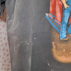 Peut inclure: T-shirt gris fonc&eacute; avec un graphisme color&eacute; d'un homme en salopette bleue et d'une structure rouge et bleue. Le t-shirt a un aspect us&eacute; avec quelques marques blanches.