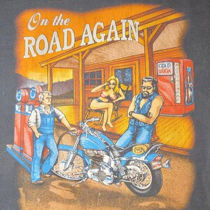 Peut inclure: T-shirt noir avec un motif repr&eacute;sentant une moto, une station-service et trois personnages. Le texte "On the Road Again" est affich&eacute; au-dessus de la sc&egrave;ne. L'&oelig;uvre d'art a un aspect vintage et l&eacute;g&egrave;rement d&eacute;lav&eacute;.