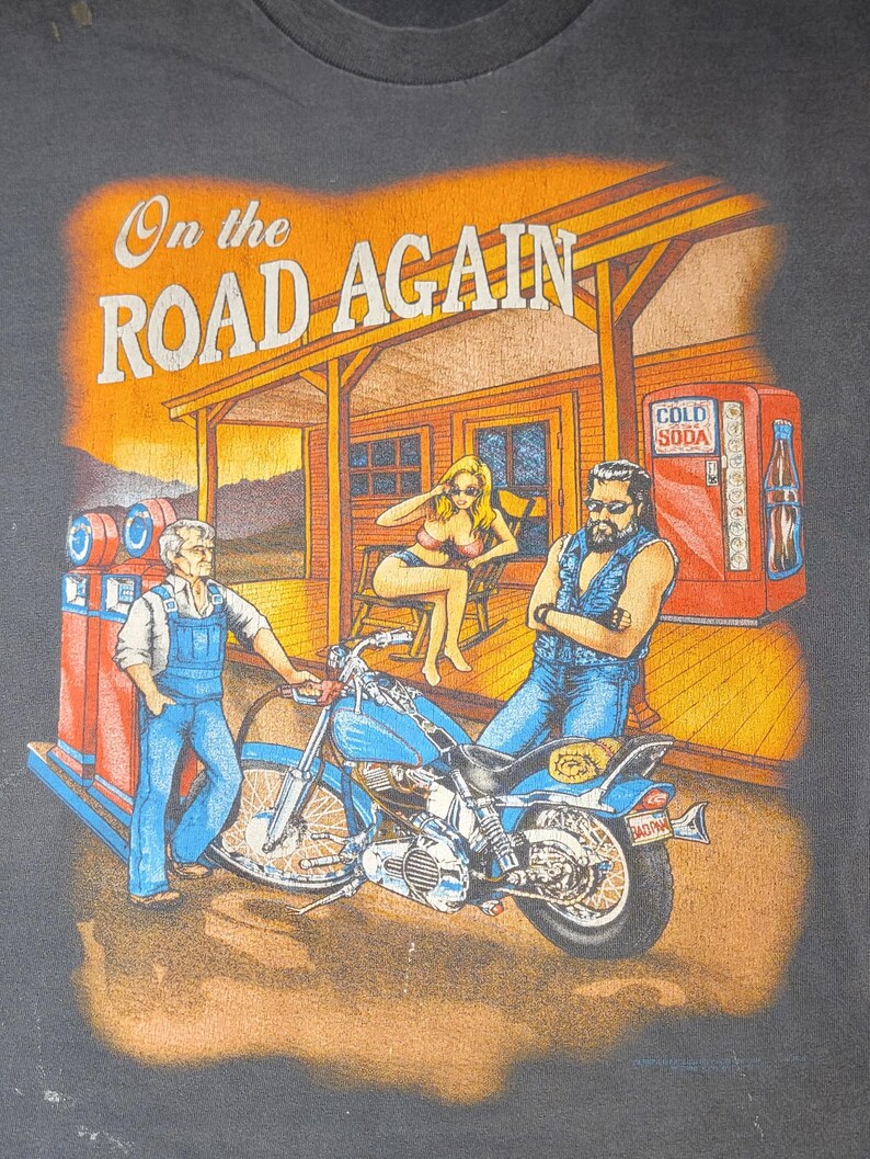 Peut inclure: T-shirt noir avec un motif repr&eacute;sentant une moto, une station-service et trois personnages. Le texte "On the Road Again" est affich&eacute; au-dessus de la sc&egrave;ne. L'&oelig;uvre d'art a un aspect vintage et l&eacute;g&egrave;rement d&eacute;lav&eacute;.