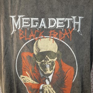 May include: Gray t-shirt featuring the band name "MEGADETH" above the words "BLACK FRIDAY" in orange. The graphic is a skeleton in a suit and sunglasses. The shirt is on a mannequin.