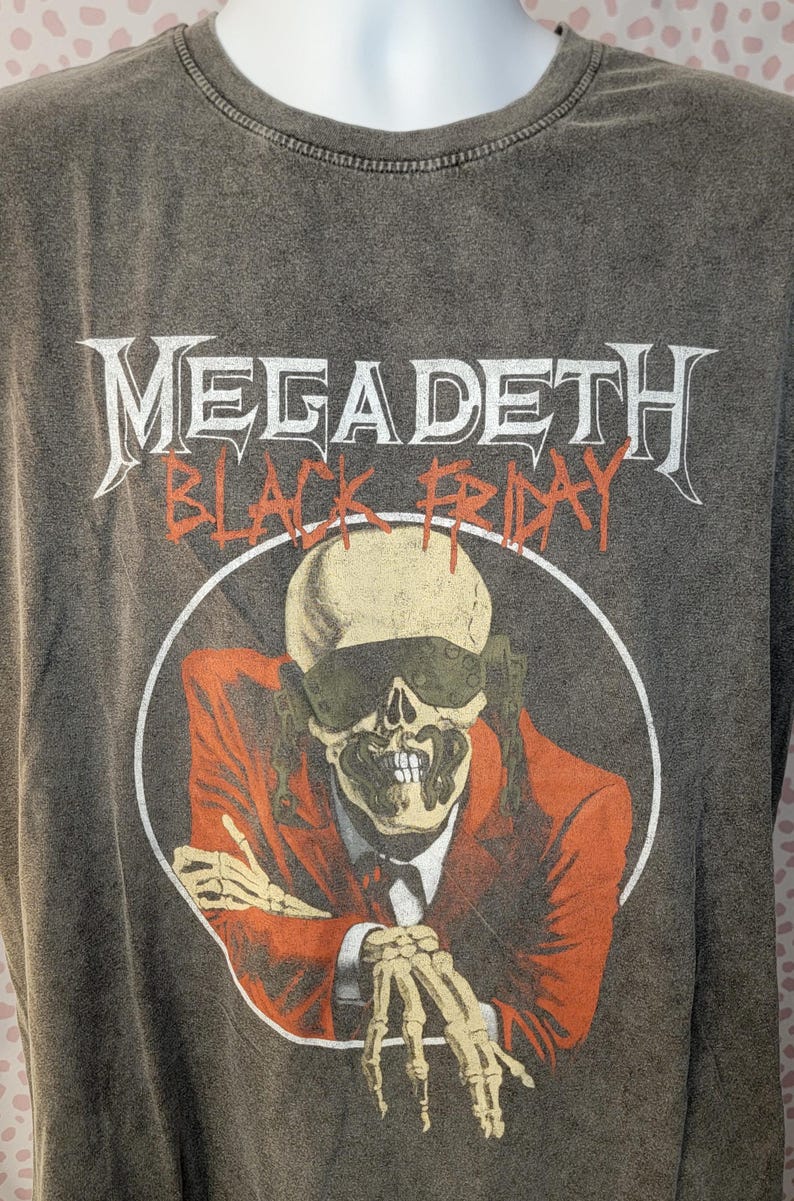 May include: Gray t-shirt featuring the band name "MEGADETH" above the words "BLACK FRIDAY" in orange. The graphic is a skeleton in a suit and sunglasses. The shirt is on a mannequin.