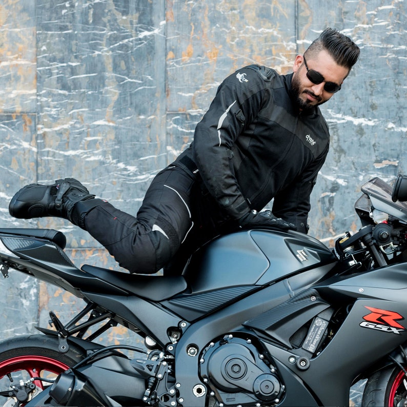 K&ouml;nnte beinhalten: Eine Person in einer schwarzen Motorradjacke und -hose sitzt auf einem schwarzen Motorrad. Das Motorrad hat einen schwarzen Sitz und einen schwarzen und silbernen Auspuff.