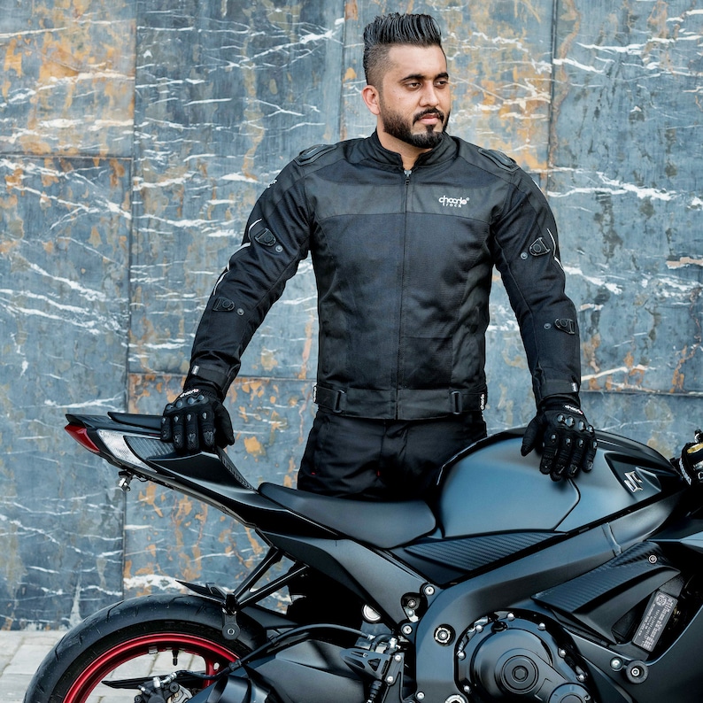 K&ouml;nnte beinhalten: Eine schwarze Motorradjacke mit einem grauen Einsatz auf der Brust und dem Wort "Choole" auf dem R&uuml;cken. Die Jacke hat reflektierende Akzente und wird von einer Person getragen, die neben einem schwarzen Motorrad steht.
