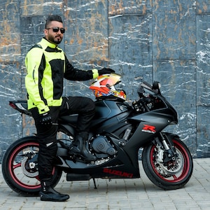K&ouml;nnte beinhalten: Eine Person in einer gelben und schwarzen Motorradjacke sitzt auf einem schwarzen Suzuki Motorrad mit einem roten und gelben Helm auf dem Soziussitz.