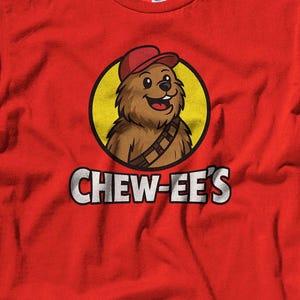 May include: A red t-shirt featuring a cartoon illustration of a furry creature wearing a red cap and a utility belt, set within a yellow circle. The text "CHEW-EE'S" is printed below the image in white letters.