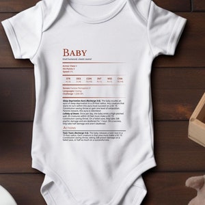 Op de afbeelding: Witte babybody met het woord "BABY" in kastanjebruine tekst. De body heeft een gedetailleerde tabel erop gedrukt. Een houten kist met een stripfiguur, een knuffelhond, een kopje koffie en houten blokken staan ook in de afbeelding.