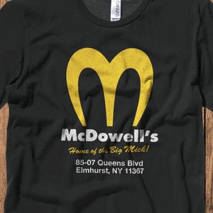 May include: Black t-shirt featuring a yellow stylized "M" logo and the text "McDowell's Home of the Big Mick!" with an address below. The shirt is made of a soft material.