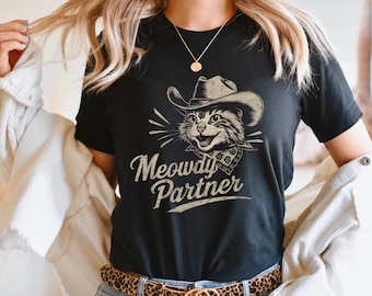 May include: Black t-shirt with a graphic of a cat wearing a cowboy hat and a bandana. The text "Meowdy Partner" is printed below the cat.