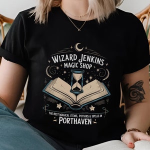 May include: Black t-shirt featuring the text "WIZARD JENKINS MAGIC SHOP" above an open book with an hourglass. The text "THE BEST MAGICAL ITEMS, POTIONS & SPELLS IN PORTHAVEN" is below the book. The design includes stars and a crescent moon.