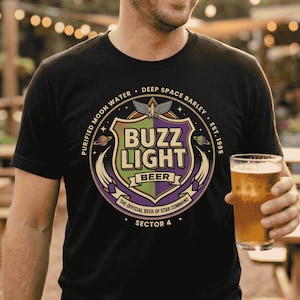 May include: Black t-shirt with a colorful graphic design. The design features the words "BUZZ LIGHT BEER" in a shield shape, with the text "PURIFIED MOON WATER" and "DEEP SPACE BARLEY" around it. A glass of beer is held in the hand.