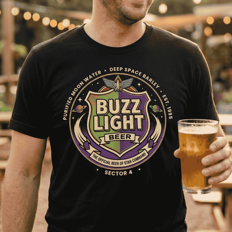 May include: Black t-shirt with a colorful graphic design. The design features the words "BUZZ LIGHT BEER" in a shield shape, with the text "PURIFIED MOON WATER" and "DEEP SPACE BARLEY" around it. A glass of beer is held in the hand.