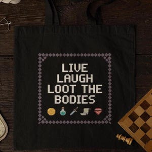 May include: Black tote bag with the text "Live Laugh Loot the Bodies" in a pixelated font. The text is surrounded by a purple border. There are five pixelated icons below the text: a gold coin, a potion bottle, a dagger, a boot, and a diamond.