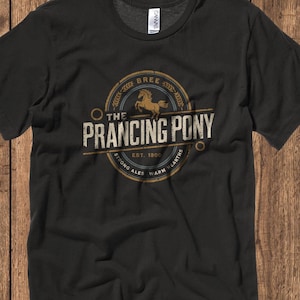 May include: Black t-shirt featuring a vintage-style logo for "The Prancing Pony" brewery. The logo depicts a brown horse, the words "The Prancing Pony", and the text "Bree Est. 1800 Strong Ales Warm Hearths".
