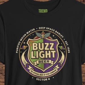 May include: A black t-shirt featuring a circular graphic with a shield design in green and purple. The text reads BUZZ LIGHT BEER, PURIFIED MOON WATER, DEEP SPACE BARLEY, EST. 1995, THE OFFICIAL BEER OF STAR COMMAND, and SECTOR 4, with a small rocket icon.