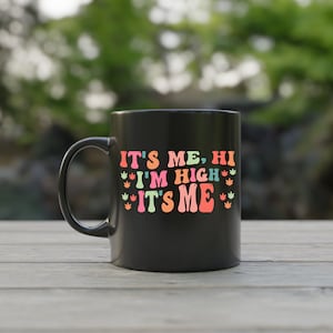 May include: Black ceramic mug with a white design that says "It's me, hi I'm high It's me" with colorful marijuana leaf graphics.