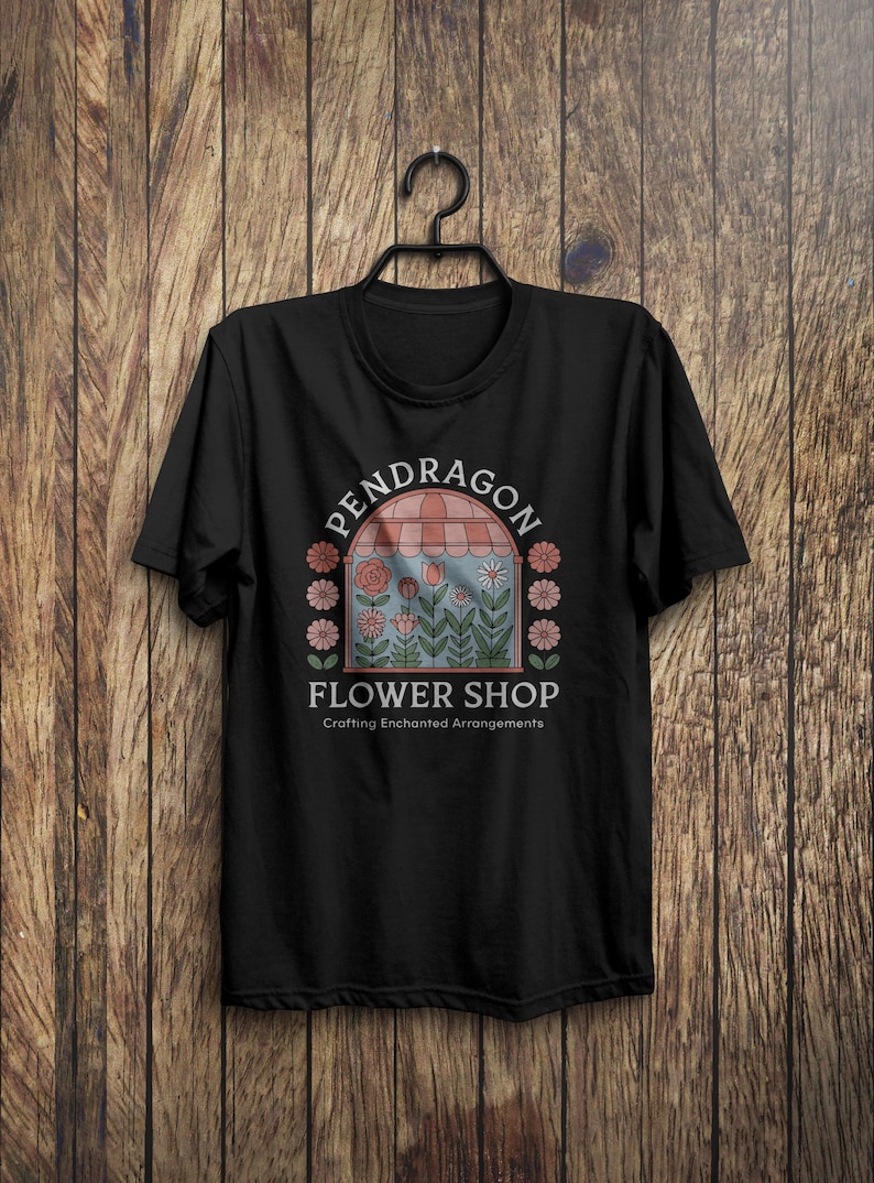 K&ouml;nnte beinhalten: Schwarzes T-Shirt mit einem Aufdruck eines Blumenladens mit dem Text "Pendragon Flower Shop" und "Crafting Enchanted Arrangements".