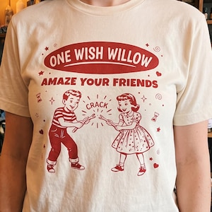 Könnte beinhalten: Ein cremefarbenes Kurzarm-T-Shirt mit rotem Grafikdruck. Der Text lautet ONE WISH WILLOW und AMAZE YOUR FRIENDS. Die Illustration zeigt einen Jungen und ein Mädchen, die an einem Wunschknochen ziehen, mit dem Wort CRACK dazwischen.