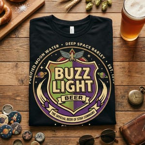 May include: Black t-shirt featuring a shield design with the words "BUZZ LIGHT BEER" in green and purple. The design includes the text "PURIFIED MOON WATER" and "DEEP SPACE BARLEY". The shirt also has the text "THE OFFICIAL BEER OF STAR COMMAND".