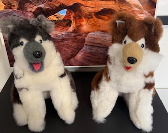 Op de afbeelding: Twee pluche hondenspeeltjes. Eén is grijs, zwart en wit, met blauwe ogen en een roze tong. De andere is bruin, wit en beige, met blauwe ogen en een rode tong. Beide zitten.