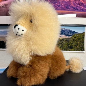 Op de afbeelding: Een pluche leeuwenknuffel met een lichtbruin lichaam en een pluche, cr&egrave;mekleurige manen. De leeuw heeft een zwarte neus en zwarte ogen.