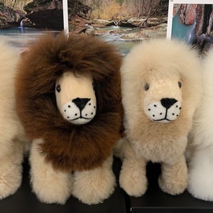Op de afbeelding: Vier pluche leeuwen speelgoed in verschillende kleuren: wit, bruin, beige en wit. De leeuwen hebben zwarte ogen en neuzen en zijn gemaakt van zacht, pluche materiaal.