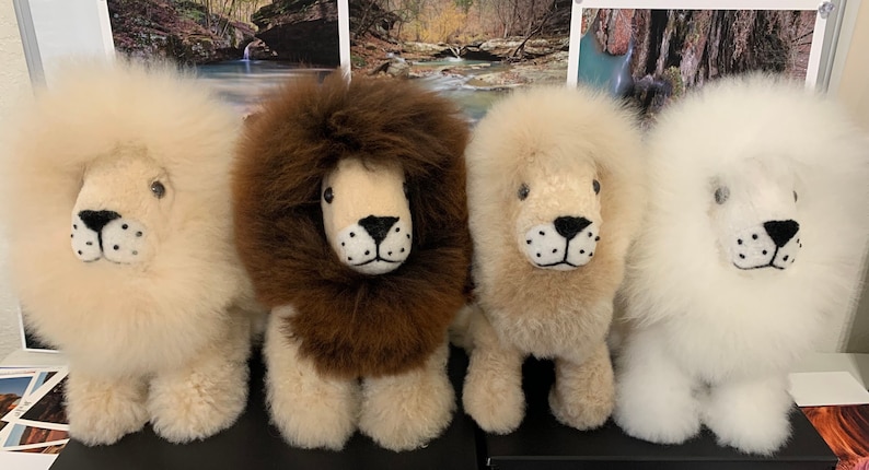 Op de afbeelding: Vier pluche leeuwen speelgoed in verschillende kleuren: wit, bruin, beige en wit. De leeuwen hebben zwarte ogen en neuzen en zijn gemaakt van zacht, pluche materiaal.