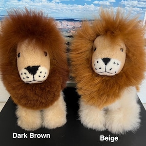 Op de afbeelding: Twee pluche leeuwen speelgoed, een met donkerbruine vacht en de andere met beige vacht. Beide hebben een wit gezicht met zwarte ogen en een zwarte neus.