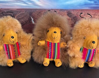 Op de afbeelding: Drie pluche alpaca speelgoedjes met bruin haar en kleurrijke gestreepte stoffen accenten. De speelgoedjes staan op een zwart oppervlak.